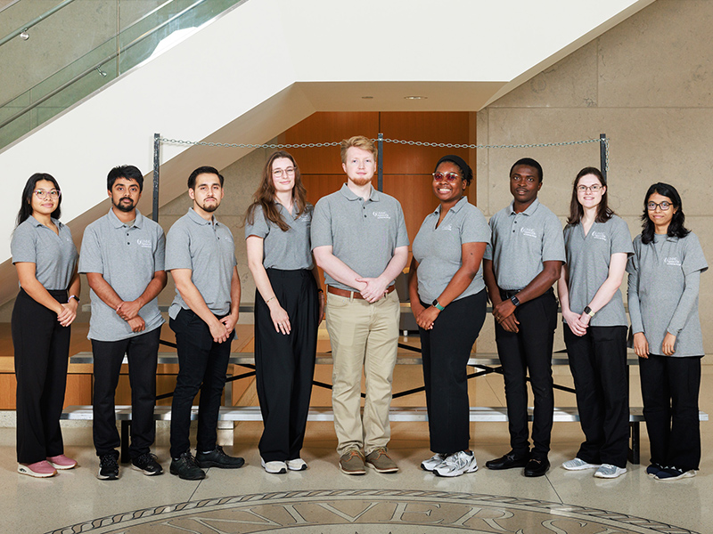 Group portrait of nine Microbiology and Immunology students in the lobby of the School of Medicine. A staircase is behind them.